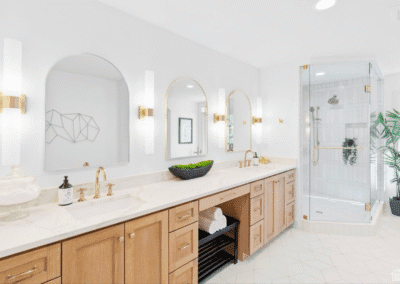 Modern bathroom with double sinks, wooden cabinets, large mirrors, wall sconces, and a glass-enclosed shower. Decorative plants and geometric wall art add style.