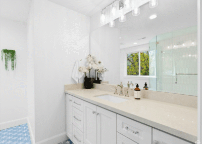 Modern bathroom with white cabinets, beige countertop, large mirror, glass shower, decorative plants, and blue patterned floor tiles.